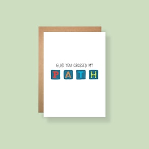 May include: A white greeting card with a brown envelope featuring the black text GLAD YOU CROSSED MY and the word PATH below it, where each letter is inside a blue square with distinct colors: red P, orange A, light blue T, and yellow H.