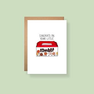 May include: A white greeting card featuring a red and white box illustration with the text CONGRATS ON YOUR LITTLE timbit. The box contains several small round doughnut holes with drawn legs and feet, appearing to walk out of the open container.