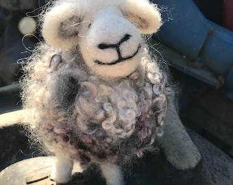 May include: A white and gray wool felt sheep with a black nose and mouth. The sheep is standing on a brown surface.