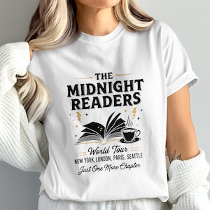 Può includere: T-shirt bianca con un design grafico nero. Il design include il testo "THE MIDNIGHT READERS" sopra un libro aperto, una tazza di caffè e le parole "World Tour" e "Just One More Chapter".