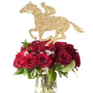 May include: A gold glitter horse and rider silhouette centerpiece for a wedding or party. The horse is running and the rider is wearing a jockey cap. The centerpiece is placed on top of a bouquet of red roses in a clear glass vase.