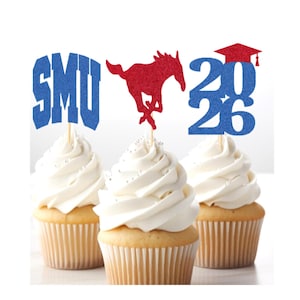 May include: Three cupcakes with white frosting, topped with blue and red glitter cake toppers. The toppers spell out "SMU", a red mustang, and the year "2026" with a graduation cap. The cupcakes are in tan paper liners.