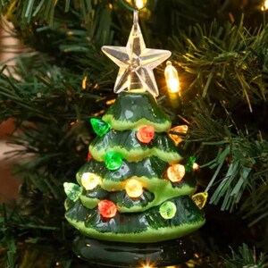 May include: A ceramic Christmas tree ornament with a clear star topper. The green tree is adorned with colorful lights in red, green, and yellow. The ornament is set against a backdrop of a green Christmas tree.
