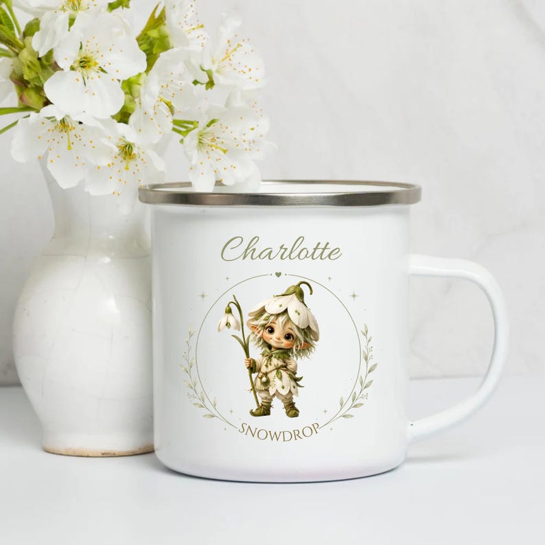 Könnte beinhalten: Weißer Emaillebecher mit Silberrand. Er zeigt eine Schneeglöckchen-Feen-Illustration, die eine Blume hält, mit dem Namen "Charlotte" und dem Wort "Snowdrop". Im Hintergrund steht eine Vase mit weißen Blumen. Ein bezauberndes Geschenk.