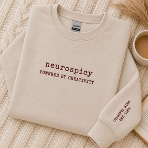 Könnte beinhalten: Ein sandfarbenes Sweatshirt mit Rundhalsausschnitt und dunkelroter Stickerei auf der Brust mit dem Text "neurospicy POWERED BY CREATIVITY". Am linken Ärmelbündchen befindet sich eine weitere dunkelrote Stickerei "COLORFUL MIND EST. 1984".