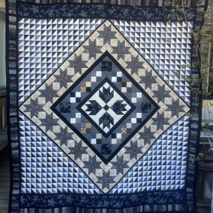 Op de afbeelding: Een zwarte, witte en grijze quilt met een geometrisch patroon. De quilt is gemaakt van driehoeken, sterren en vierkanten. De quilt hangt aan een waslijn.