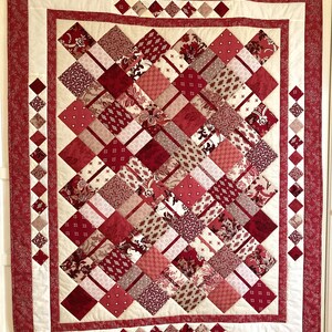 Op de afbeelding: Een rood en wit quilt met een ruitpatroon. De quilt is gemaakt van verschillende stoffen met verschillende patronen. De quilt hangt aan een muur.