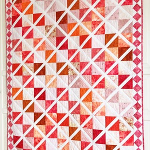 May include: A quilt with a geometric pattern of pink, orange, and white triangles. The quilt is edged with a pink and white diamond pattern.