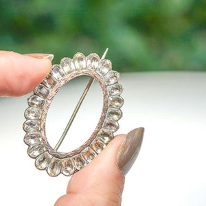 Op de afbeelding: Een ovale broche met een zilverkleurig metalen frame en heldere, facetgeslepen stenen. De broche heeft een speld op de achterkant om hem aan kleding te bevestigen. De stenen zijn rond het ovaal gerangschikt. Sieraden.