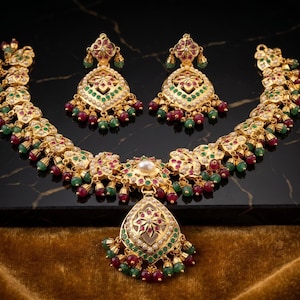 May include: A gold-toned necklace and matching earrings set. The jewelry features intricate designs with red, green, and white gemstones. The necklace has a teardrop pendant. The set is displayed on a black surface.