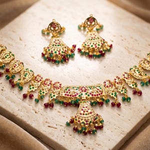 May include: A gold-toned jewelry set featuring a necklace and matching earrings. The necklace and earrings are adorned with red and green gemstones. The jewelry is displayed on a light-colored, textured surface.