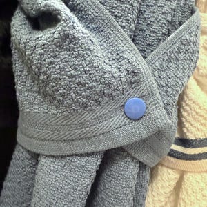 May include: A blue textured hanging kitchen towel featuring a folded top section secured with a single round blue plastic snap button. The towel has a woven fabric texture and a decorative border along the folded edge, hanging vertically.