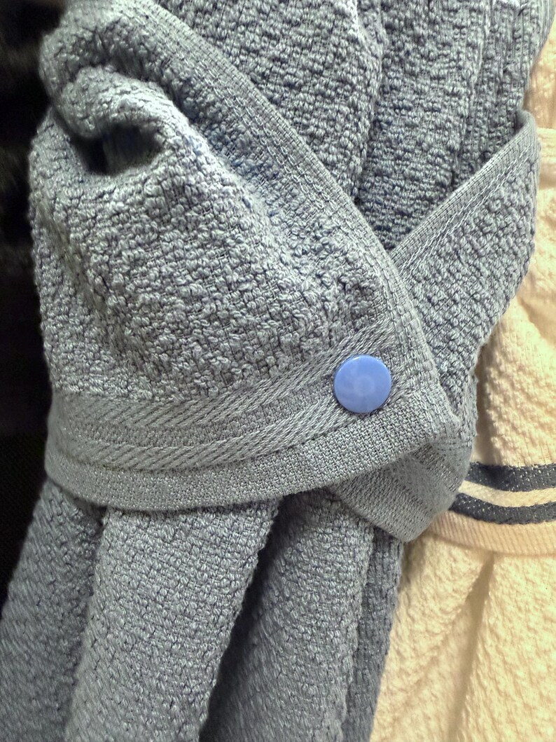 May include: A blue textured hanging kitchen towel featuring a folded top section secured with a single round blue plastic snap button. The towel has a woven fabric texture and a decorative border along the folded edge, hanging vertically.
