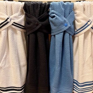 May include: Four hanging kitchen towels with a textured waffle weave pattern. Two towels are white with black or blue striped accents, one is solid black, and one is solid blue. Each towel features a top loop secured with a matching plastic snap button.