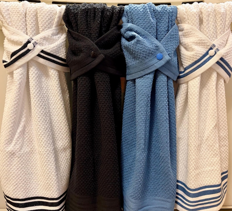 May include: Four hanging kitchen towels with a textured waffle weave pattern. Two towels are white with black or blue striped accents, one is solid black, and one is solid blue. Each towel features a top loop secured with a matching plastic snap button.