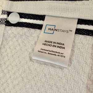May include: A white terry cloth towel featuring two horizontal navy blue stripes and a white circular button. A white fabric tag is attached, displaying the text Mainstays, Made in India, Hecho en India, and Distributed by Walmart Inc., Bentonville, AR 72716.