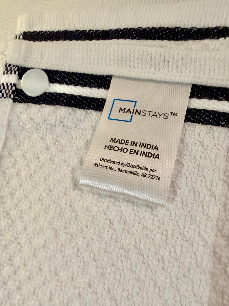 May include: A white terry cloth towel featuring two horizontal navy blue stripes and a white circular button. A white fabric tag is attached, displaying the text Mainstays, Made in India, Hecho en India, and Distributed by Walmart Inc., Bentonville, AR 72716.