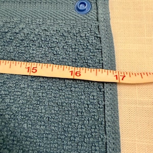 May include: A blue terry cloth towel featuring a single blue plastic snap fastener at the top corner. A white measuring tape is positioned horizontally across the fabric, showing a width measurement extending past the 16-inch mark.