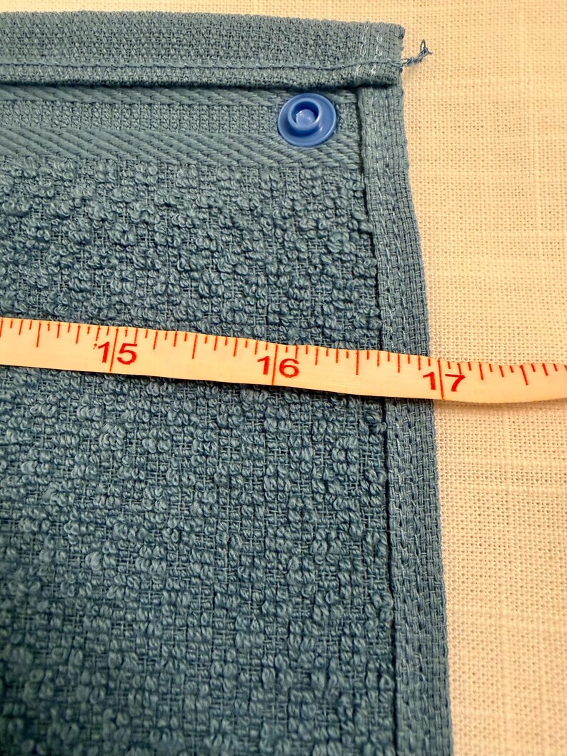 May include: A blue terry cloth towel featuring a single blue plastic snap fastener at the top corner. A white measuring tape is positioned horizontally across the fabric, showing a width measurement extending past the 16-inch mark.