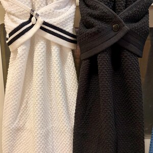 May include: Two hanging kitchen towels with a textured waffle weave pattern. One towel is white with black striped trim and a white button, while the other is solid charcoal gray with a matching dark button. Both towels feature a folded top loop.