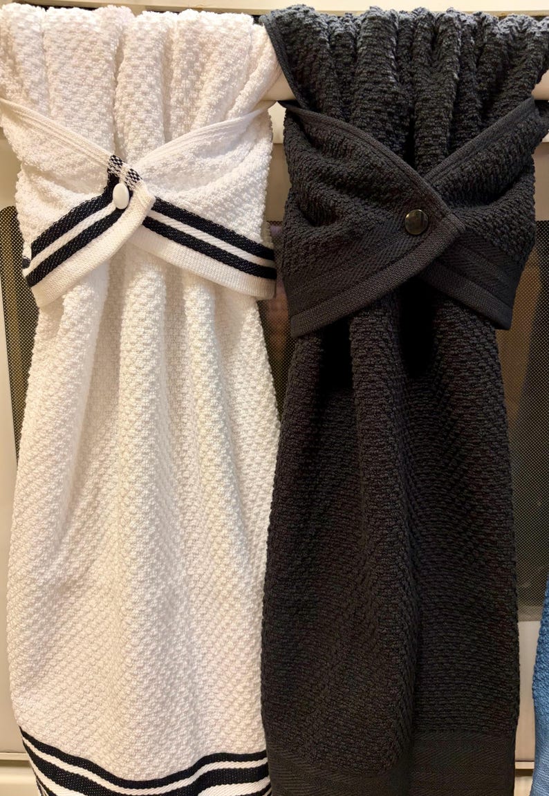 May include: Two hanging kitchen towels with a textured waffle weave pattern. One towel is white with black striped trim and a white button, while the other is solid charcoal gray with a matching dark button. Both towels feature a folded top loop.
