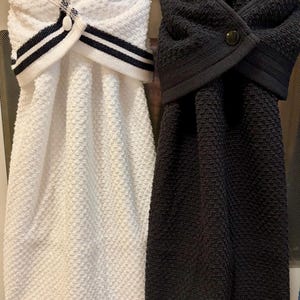 May include: Two hanging kitchen towels with a textured waffle weave pattern. One towel is white with black striped trim and a white button, while the other is solid black with a matching black button. Both feature a folded top closure.