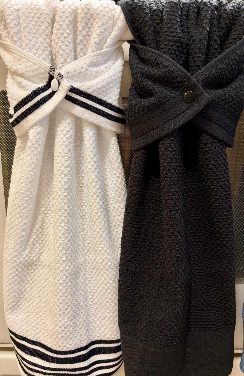 May include: Two hanging kitchen towels with a textured waffle weave pattern. One towel is white with black striped trim and a white button, while the other is solid black with a matching black button. Both feature a folded top closure.