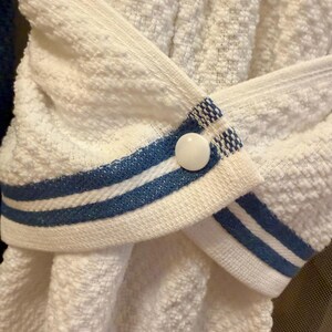 May include: A white textured kitchen towel featuring a hanging loop with two horizontal blue stripes and a single white plastic snap button closure. The towel is folded and secured in the center by the buttoned loop, creating a gathered appearance.