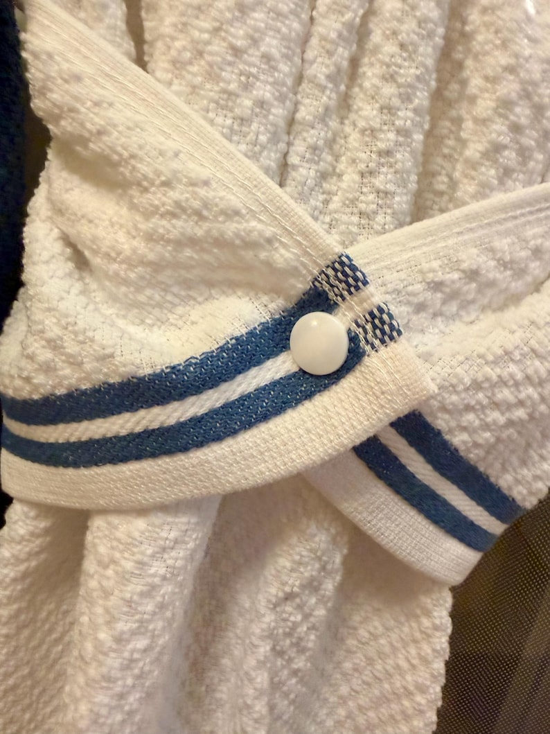 May include: A white textured kitchen towel featuring a hanging loop with two horizontal blue stripes and a single white plastic snap button closure. The towel is folded and secured in the center by the buttoned loop, creating a gathered appearance.