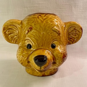May include: A ceramic bear head with a glossy finish. The bear has a brown nose, black eyes, and large ears. The head is a light brown color with darker brown accents. The bear head is likely a decorative item or container.