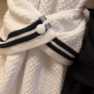 May include: A white textured kitchen towel featuring a decorative strap with two black horizontal stripes. The strap is secured in the center by a single white circular button, creating a folded, overlapping design across the fabric.