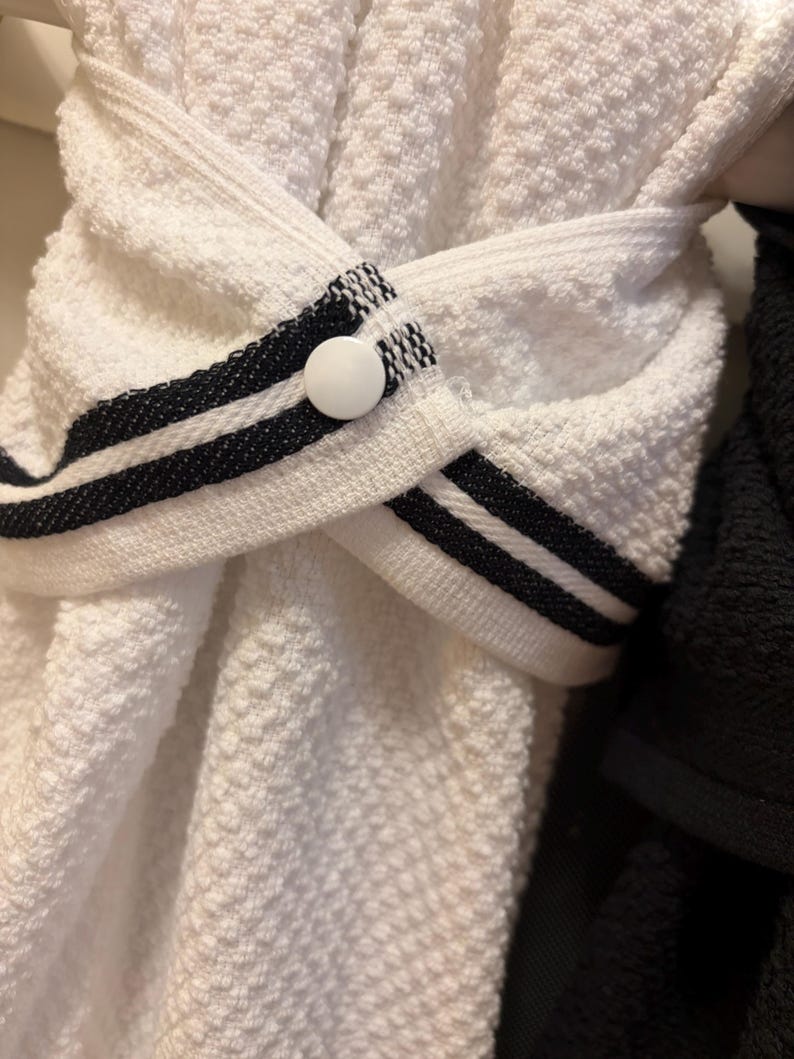 May include: A white textured kitchen towel featuring a decorative strap with two black horizontal stripes. The strap is secured in the center by a single white circular button, creating a folded, overlapping design across the fabric.