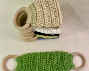 May include: A stack of wooden rings with a beige crocheted cover, and a green crocheted toy with wooden rings on each end. The crocheted items have a handmade appearance. The stack of rings has a few layers of different colored crochet.