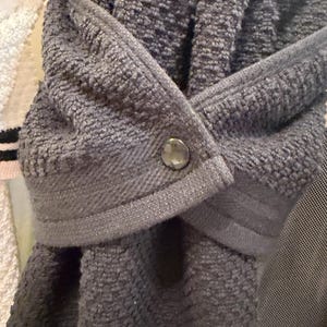 May include: A dark gray hanging kitchen towel featuring a textured terry cloth fabric and a smooth, flat-woven top band. The towel is folded over and secured in the center with a single, round, metallic-toned snap button fastener.