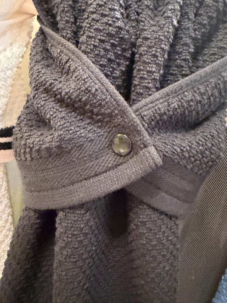 May include: A dark gray hanging kitchen towel featuring a textured terry cloth fabric and a smooth, flat-woven top band. The towel is folded over and secured in the center with a single, round, metallic-toned snap button fastener.