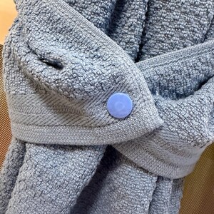 May include: A light blue textured towel featuring a woven border and a single round light blue plastic snap button closure. The fabric has a looped pile construction, and the towel is folded over itself to showcase the secure button fastening.