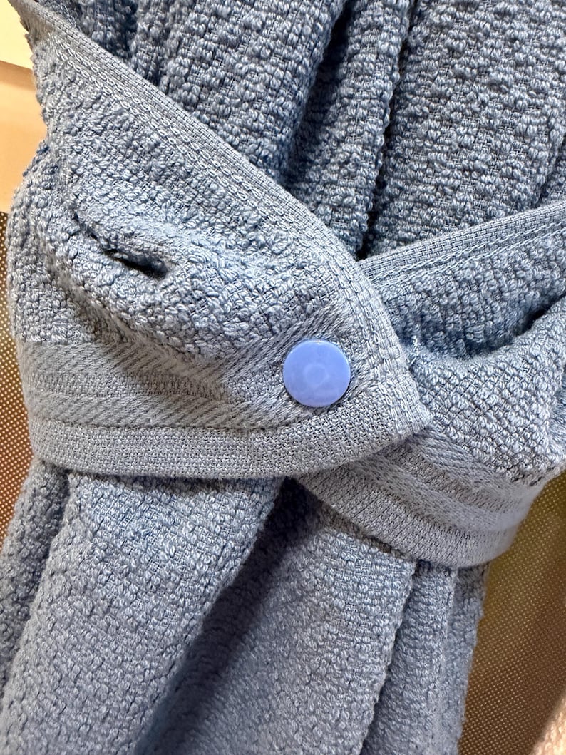 May include: A light blue textured towel featuring a woven border and a single round light blue plastic snap button closure. The fabric has a looped pile construction, and the towel is folded over itself to showcase the secure button fastening.