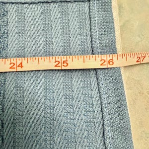 May include: A light blue fabric with a textured, vertical chevron-like weave pattern. A yellow measuring tape is positioned horizontally across the fabric, showing a measurement spanning from the 24-inch mark to the 27-inch mark.