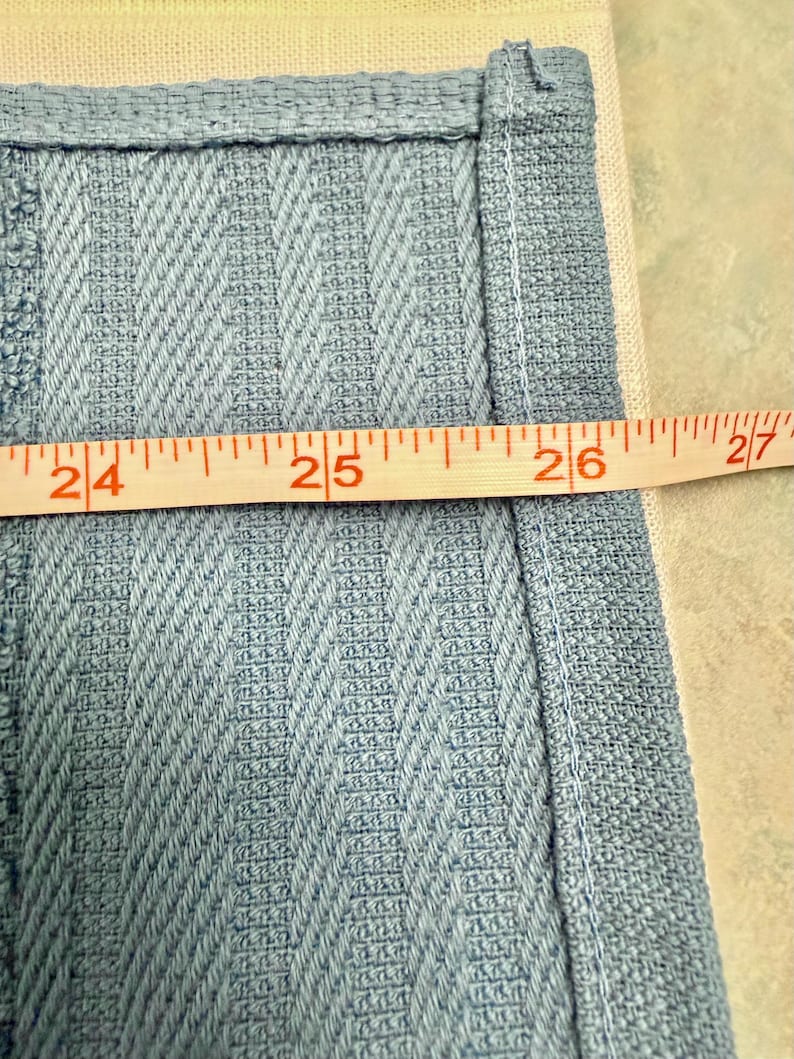 May include: A light blue fabric with a textured, vertical chevron-like weave pattern. A yellow measuring tape is positioned horizontally across the fabric, showing a measurement spanning from the 24-inch mark to the 27-inch mark.