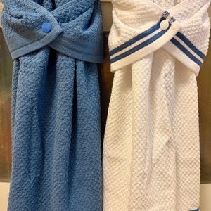 May include: Two hanging kitchen towels with a textured weave. One towel is solid blue with a blue snap button, and the other is white with blue striped trim and a white snap button. Both towels feature a folded top section that secures with a button.