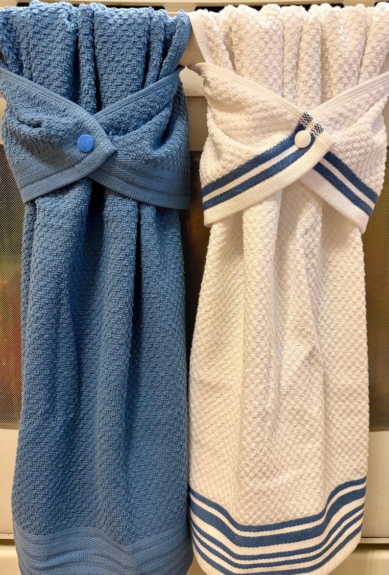May include: Two hanging kitchen towels with a textured weave. One towel is solid blue with a blue snap button, and the other is white with blue striped trim and a white snap button. Both towels feature a folded top section that secures with a button.