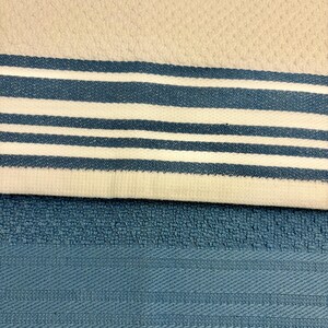 May include: A kitchen towel with a textured off-white surface featuring a horizontal band of three blue stripes and two white stripes, layered over a solid blue kitchen towel with a textured weave and a subtle horizontal striped pattern.