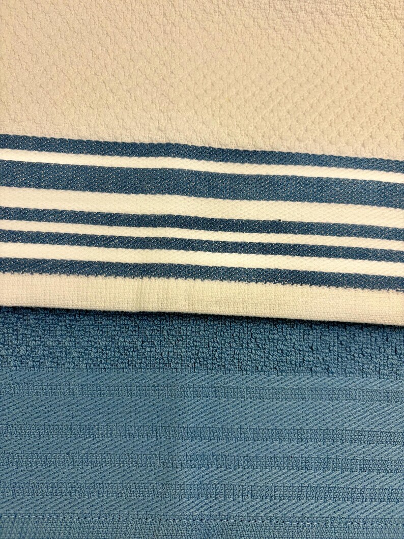 May include: A kitchen towel with a textured off-white surface featuring a horizontal band of three blue stripes and two white stripes, layered over a solid blue kitchen towel with a textured weave and a subtle horizontal striped pattern.