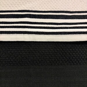 May include: A textured kitchen towel featuring a cream-colored section with a pattern of black horizontal stripes, positioned above a solid black section with a subtle woven herringbone border detail. The fabric has a distinct, raised waffle-like weave.