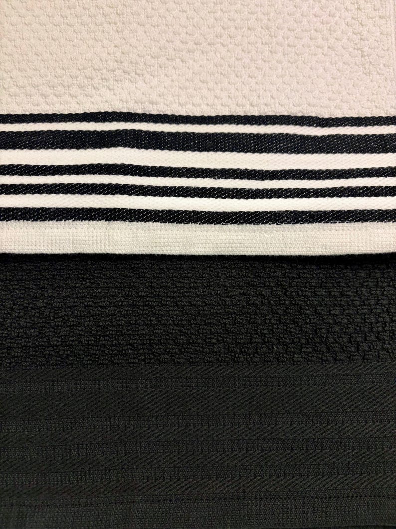 May include: A textured kitchen towel featuring a cream-colored section with a pattern of black horizontal stripes, positioned above a solid black section with a subtle woven herringbone border detail. The fabric has a distinct, raised waffle-like weave.