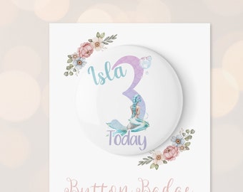 Puede incluir: Una insignia de botón blanca con una ilustración de sirena morada y azul y el texto "Isla 3 Today". La insignia está rodeada de un diseño floral con flores rosas, blancas y verdes. El texto "Button Badge" está escrito en cursiva rosa debajo de la insignia. El texto "This item is not a toy, due to the sharp pin children must be supervised" está escrito en negro debajo de la insignia. El texto "www.etsy.com/shop/littlelouscrafts" está escrito en negro debajo del texto.