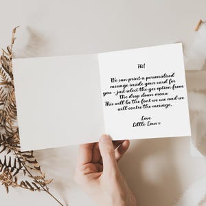 May include: Open greeting card with a handwritten message inside. The card is held open, revealing text in black script. Dried fern and a white ribbon are arranged around the card.