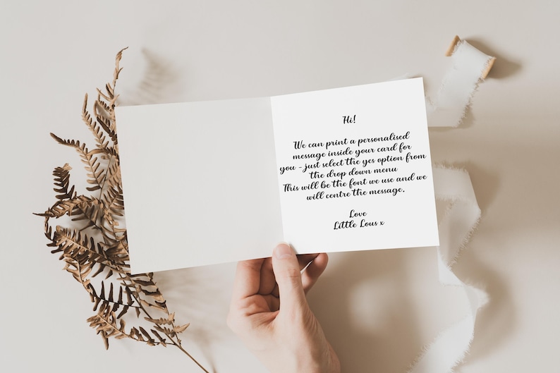 May include: Open greeting card with a handwritten message inside. The card is held open, revealing text in black script. Dried fern and a white ribbon are arranged around the card.