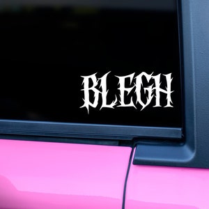 May include: A white vinyl sticker with the word "BLEGH" in a gothic font on a black background. The sticker is on the rear window of a pink car.