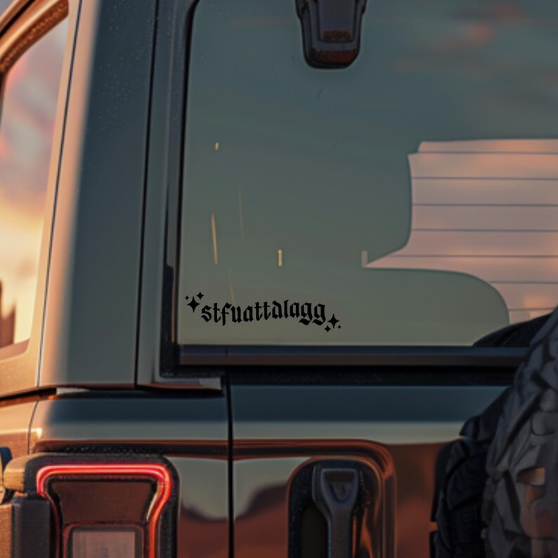 May include: Black vinyl decal with the text "*stfuattalagg*" in a gothic font, applied to the rear window of a dark-coloured vehicle. The decal is positioned on the lower right side of the window.